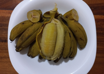 Steamed Cambodian Chek Kramuon wax bananas arranged on a white plate over a wooden table, one banana peeled open to show its soft golden flesh.