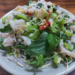 Cambodian raw fish salad on a white plate, topped with thin slices of fish, bitter melon, bean sprouts, fresh basil and mint, red chili, and lime dressing, served on a wooden table.
