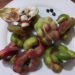 Close-up of fresh ampil teuk (Pithecellobium dulce) pods on a white plate, showing twisted green and pink Manila tamarind shells, exposed white pulp, and a few glossy black seeds, photographed on a wooden table in Cambodia.