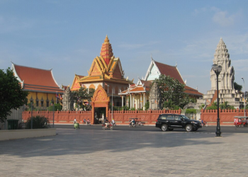 Wat Ounalom Buddhist temple complex with ornate pagodas and stupas seen from across the open square on Sisowath Quay in central Phnom Penh, Cambodia, on a sunny day with light traffic in front.