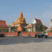 Wat Ounalom Buddhist temple complex with ornate pagodas and stupas seen from across the open square on Sisowath Quay in central Phnom Penh, Cambodia, on a sunny day with light traffic in front.