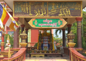 Colorful outdoor altar at Wat Sophy, Takeo, in Cambodia, decorated with Buddha images, fruits, candles, and offerings prepared to welcome the 2026 Khmer New Year angel beneath golden reliefs of the Buddha’s life.