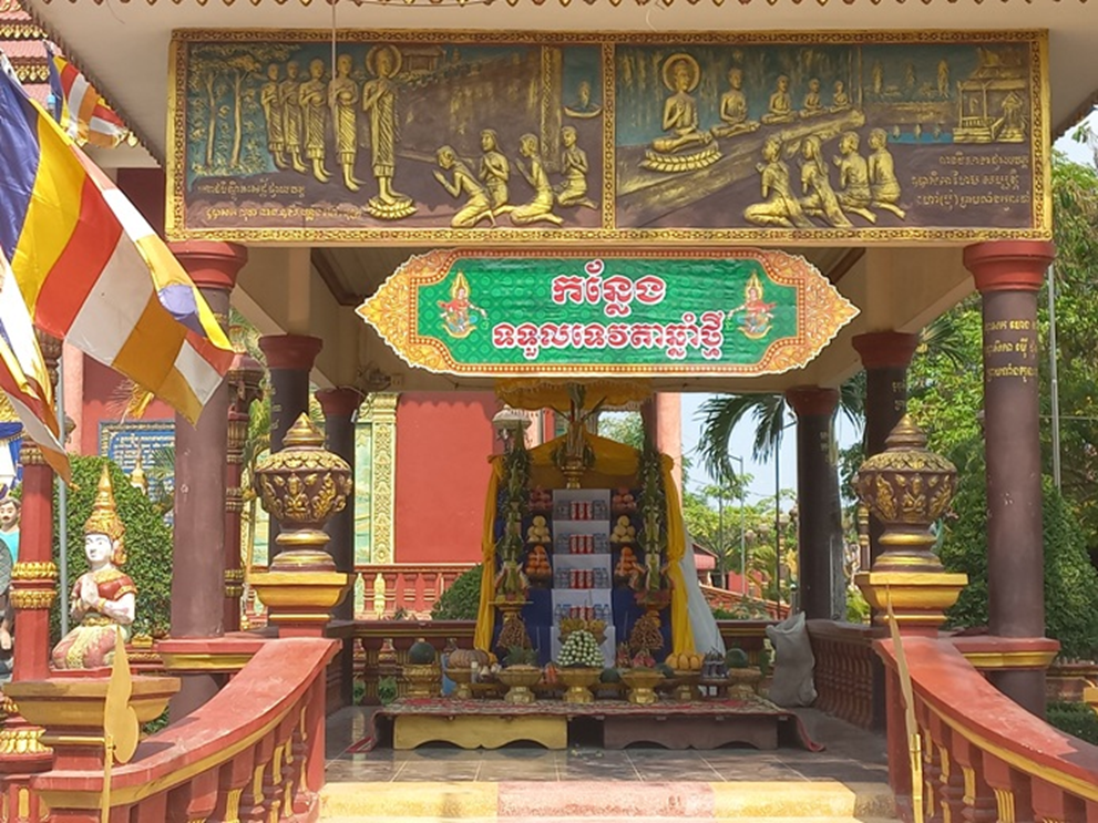 Colorful outdoor altar at Wat Sophy, Takeo, in Cambodia, decorated with Buddha images, fruits, candles, and offerings prepared to welcome the 2026 Khmer New Year angel beneath golden reliefs of the Buddha’s life.