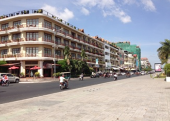 Wide riverside boulevard with a row of mid‑rise hotels and cafés on the left, scooters and cars driving along the sunlit street, and an open paved promenade with palm trees under a bright blue sky.