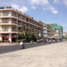 Wide riverside boulevard with a row of mid‑rise hotels and cafés on the left, scooters and cars driving along the sunlit street, and an open paved promenade with palm trees under a bright blue sky.