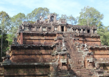 Ancient sandstone pyramid-temple of Phimeanakas in Angkor Thom, Cambodia, with steep central stairway, weathered carvings and guardian statues, rising above the jungle trees under a bright blue sky.