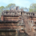 Ancient sandstone pyramid-temple of Phimeanakas in Angkor Thom, Cambodia, with steep central stairway, weathered carvings and guardian statues, rising above the jungle trees under a bright blue sky.
