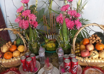 Colorful Khmer New Year family altar with fruit baskets, pink flowers, incense, sticky rice cakes, and soda cans arranged on a patterned table to welcome the New Year angel.