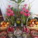 Colorful Khmer New Year family altar with fruit baskets, pink flowers, incense, sticky rice cakes, and soda cans arranged on a patterned table to welcome the New Year angel.