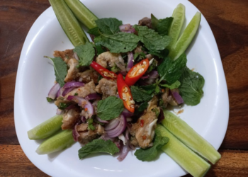 Lao-origin Cambodian deep-fried pork larb salad on a white plate, with cucumber spears, crispy pork pieces, red onion, fresh mint leaves, sliced red chilies, and a dusting of ground roasted rice.