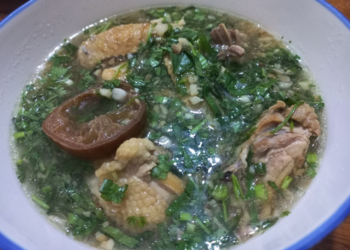 Close-up of a bowl of Cambodian ngam ngov chicken soup with preserved lime, tender chicken pieces, and chopped fresh herbs floating in a clear, aromatic broth.