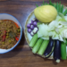 Top‑down view of Cambodian prahok ktis dip in a bowl beside a platter of fresh vegetables including cucumbers, eggplants, small purple eggplants, cabbage leaves, okra and a yellow mango on a wooden table.