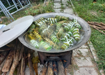 Traditional Cambodian num ansam sticky rice cakes wrapped in banana leaves steaming in outdoor cauldron over wood fire during Pchum Ben festival in countryside near Phnom Penh