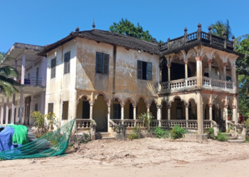 Abandoned two-story French colonial villa with arched verandas and peeling yellow walls under a bright blue sky in Angkor Borei, Takeo province, Cambodia.