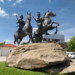 Bronze equestrian statue of Techo Meas and Techo Yat on a large rock beside the riverside promenade in Phnom Penh, with blue sky, trees and parked vehicles in the background