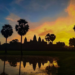 Angkor Wat temple silhouetted against a dramatic orange and yellow sunrise sky with sugar palm trees reflected in the water pool at Siem Reap, Cambodia