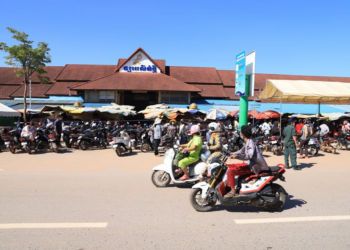 A busy street scene in front of Phsar Leu Market in Siem Reap, Cambodia, with rows of parked motorbikes, people riding scooters and walking along the road, and the red-roofed market building and vendor stalls under umbrellas in the background under a clear blue sky.
