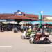 A busy street scene in front of Phsar Leu Market in Siem Reap, Cambodia, with rows of parked motorbikes, people riding scooters and walking along the road, and the red-roofed market building and vendor stalls under umbrellas in the background under a clear blue sky.