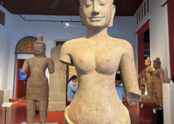 Angkor-era Bayon-style Prajnaparamita sandstone statue displayed in the National Museum of Cambodia after its 2024 return from the Denver Art Museum, with visitors viewing other Khmer sculptures in the gallery.