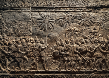 Stone bas-relief of Khmer soldiers and porters marching under palm trees, carrying weapons, drums, and baskets of rice.