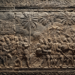 Stone bas-relief of Khmer soldiers and porters marching under palm trees, carrying weapons, drums, and baskets of rice.