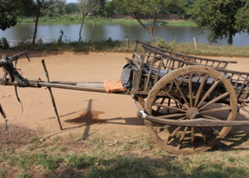 Traditional wooden Cambodian ox cart standing empty on a dirt road beside a pond in rural Banteay Meanchey, showing faded everyday transport now used mainly for tourism and cultural display.