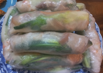 Close-up of stacked Cambodian fresh spring rolls (kuong) on a patterned blue plate, showing translucent rice paper wrapped around green herbs, pork, and shrimp.