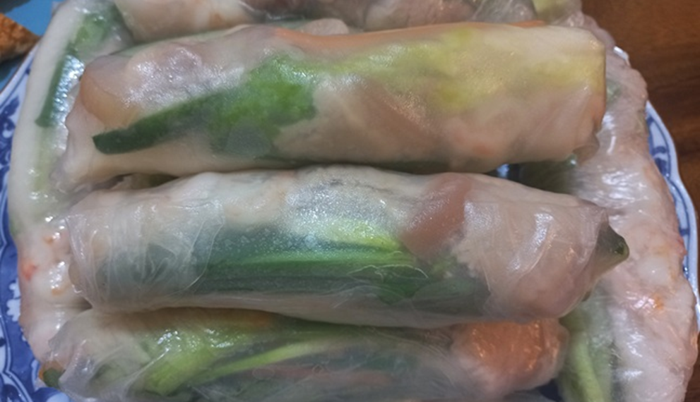 Close-up of stacked Cambodian fresh spring rolls (kuong) on a patterned blue plate, showing translucent rice paper wrapped around green herbs, pork, and shrimp.