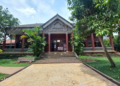 Wide-angle view of the Center for Khmer Studies library entrance at Wat Bo in Siem Reap, with red columns, carved pediment, and lush tropical trees under a bright sky.