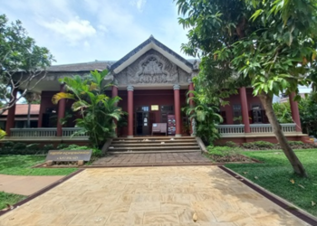 Wide-angle view of the Center for Khmer Studies library entrance at Wat Bo in Siem Reap, with red columns, carved pediment, and lush tropical trees under a bright sky.