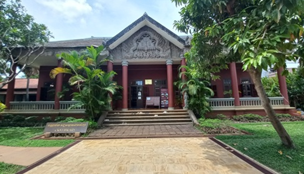 Wide-angle view of the Center for Khmer Studies library entrance at Wat Bo in Siem Reap, with red columns, carved pediment, and lush tropical trees under a bright sky.