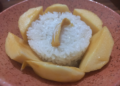 Close-up of Cambodian mango sticky rice served in a rustic clay bowl, with a round mound of glossy coconut sticky rice surrounded by golden mango slices like petals and a small jackfruit strip on top.