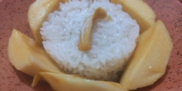 Close-up of Cambodian mango sticky rice served in a rustic clay bowl, with a round mound of glossy coconut sticky rice surrounded by golden mango slices like petals and a small jackfruit strip on top.