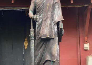 Statue of a robed Buddhist monk standing on a pedestal in front of a dark doorway and red wall, holding a closed umbrella and a book