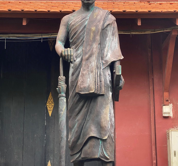 Statue of a robed Buddhist monk standing on a pedestal in front of a dark doorway and red wall, holding a closed umbrella and a book