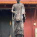 Statue of a robed Buddhist monk standing on a pedestal in front of a dark doorway and red wall, holding a closed umbrella and a book