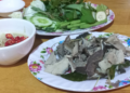 Close-up of a Cambodian meal featuring sliced boiled beef tripe on a floral plate, served with a bowl of prahok fermented fish dipping sauce topped with red chilies and a separate plate of fresh green herbs and vegetables on a wooden table.