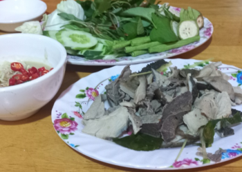 Close-up of a Cambodian meal featuring sliced boiled beef tripe on a floral plate, served with a bowl of prahok fermented fish dipping sauce topped with red chilies and a separate plate of fresh green herbs and vegetables on a wooden table.