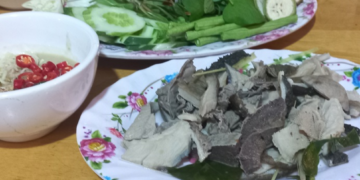 Close-up of a Cambodian meal featuring sliced boiled beef tripe on a floral plate, served with a bowl of prahok fermented fish dipping sauce topped with red chilies and a separate plate of fresh green herbs and vegetables on a wooden table.
