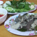 Close-up of a Cambodian meal featuring sliced boiled beef tripe on a floral plate, served with a bowl of prahok fermented fish dipping sauce topped with red chilies and a separate plate of fresh green herbs and vegetables on a wooden table.