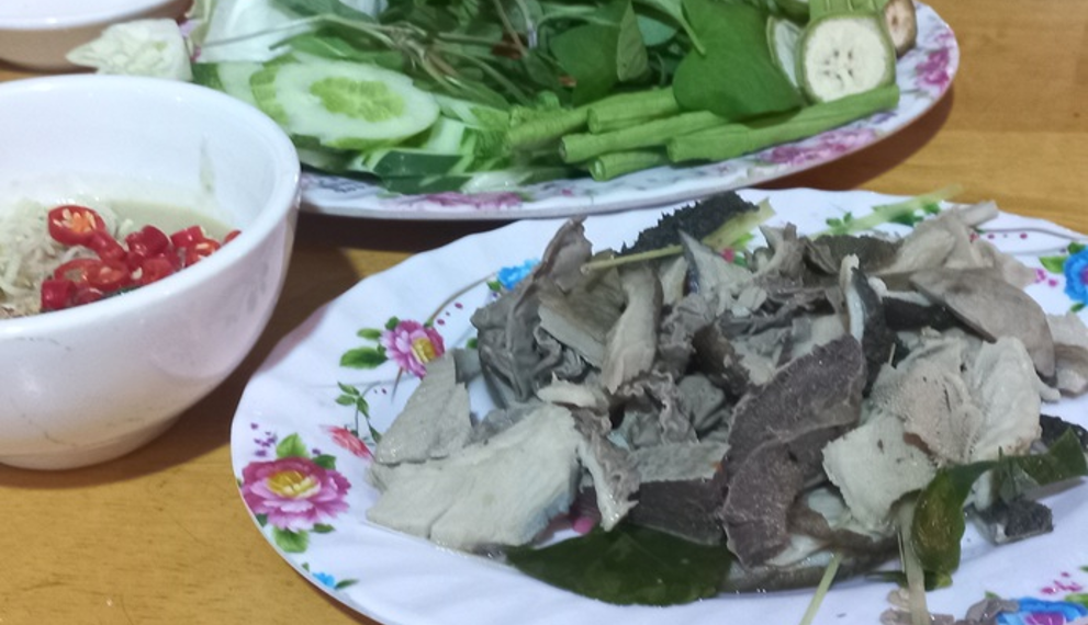 Close-up of a Cambodian meal featuring sliced boiled beef tripe on a floral plate, served with a bowl of prahok fermented fish dipping sauce topped with red chilies and a separate plate of fresh green herbs and vegetables on a wooden table.