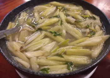 Close-up of a black bowl filled with clear Cambodian alocasia stem soup, showing pale green stems, herbs, and tiny pieces of fish in a light broth on a wooden table.