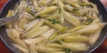 Close-up of a black bowl filled with clear Cambodian alocasia stem soup, showing pale green stems, herbs, and tiny pieces of fish in a light broth on a wooden table.