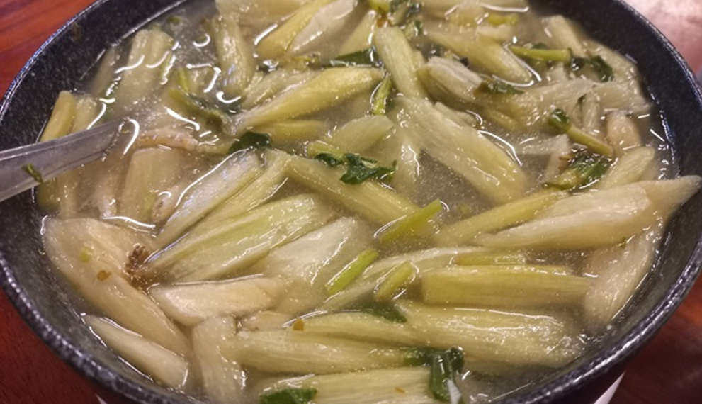Close-up of a black bowl filled with clear Cambodian alocasia stem soup, showing pale green stems, herbs, and tiny pieces of fish in a light broth on a wooden table.