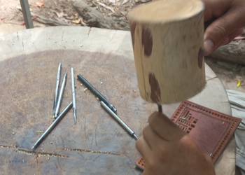 Close-up of a Siem Reap artisan using metal chisels and a wooden mallet on a leather panel to create traditional sbaek thom Khmer shadow puppets.