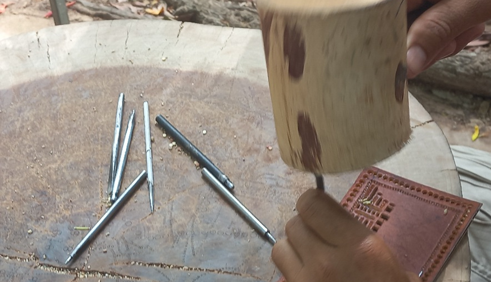 Close-up of a Siem Reap artisan using metal chisels and a wooden mallet on a leather panel to create traditional sbaek thom Khmer shadow puppets.