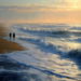 Two people walking along a wet sandy beach at dusk beside foaming waves, with golden light from the setting sun reflecting on the ocean and low clouds in the distance.