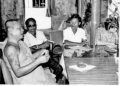 Black and white photo of monk Khieu Chum speaking at a table with three journalists in a simple indoor setting