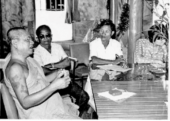 Black and white photo of monk Khieu Chum speaking at a table with three journalists in a simple indoor setting