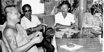 Black and white photo of monk Khieu Chum speaking at a table with three journalists in a simple indoor setting